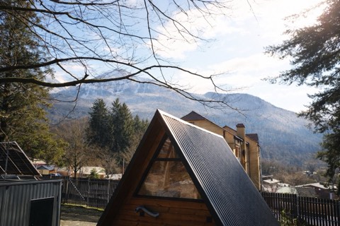 Деревянный A-frame домик с гофрированной крышей и треугольным окном на фоне заснеженных гор, Красная Поляна.